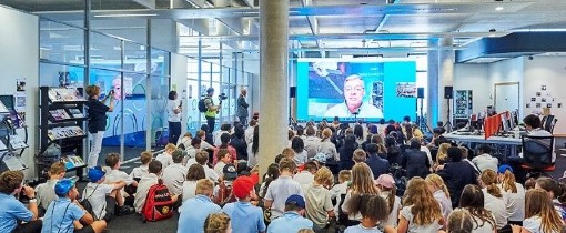 Young students sitting on the floor in a large classroom, watching a presentation on a screen
